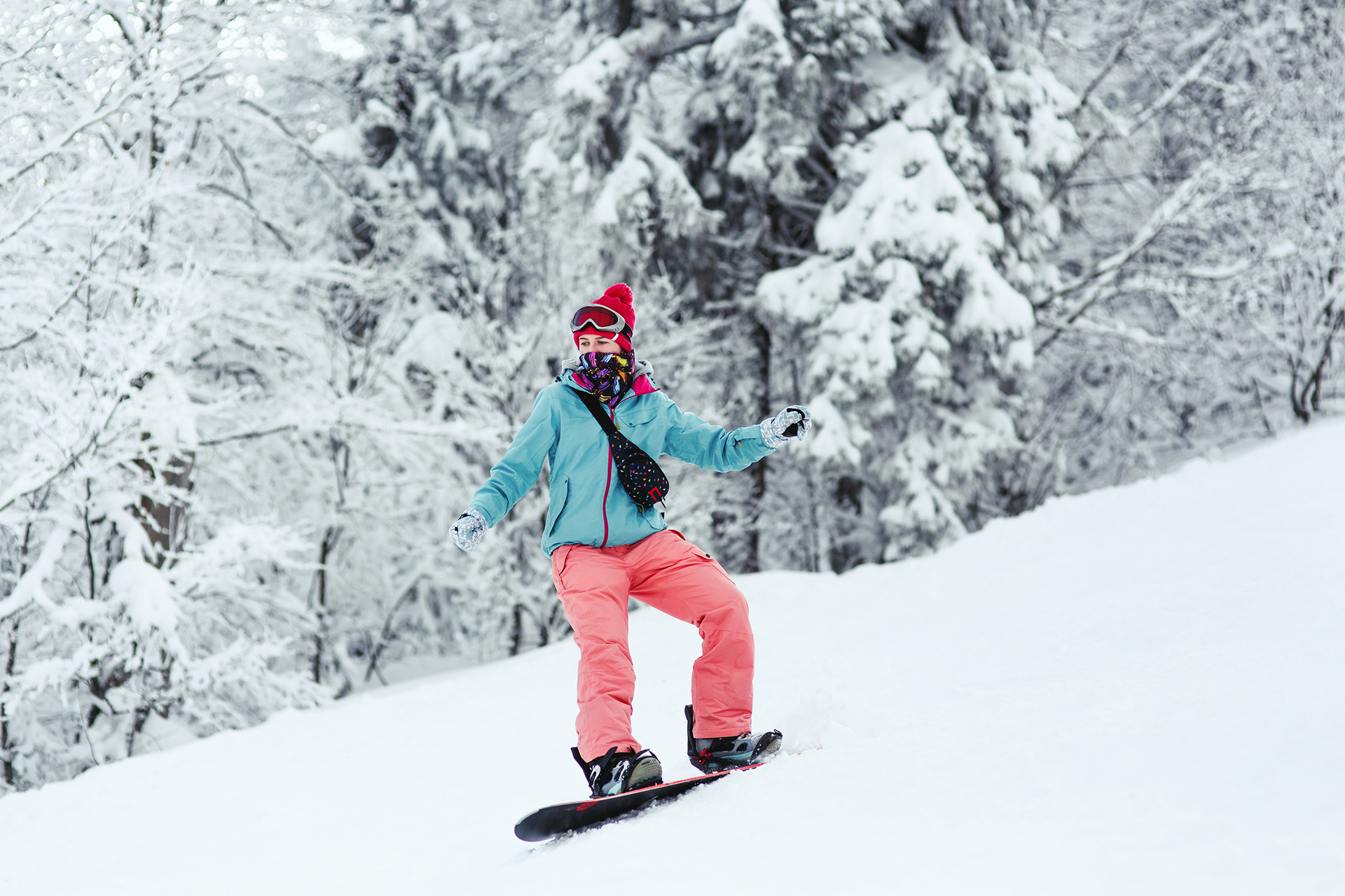 Woman in blue ski jacket and pink pants stands on the snowboard somewhere in winter forest